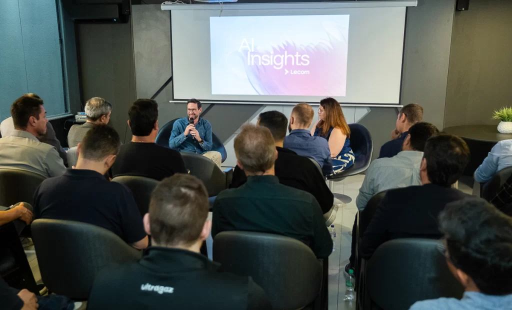 Foto tirada de trás da plateia em um auditório, mostrando um homem falando ao microfone no palco e uma tela ao fundo com a frase AI Insights Lecom.