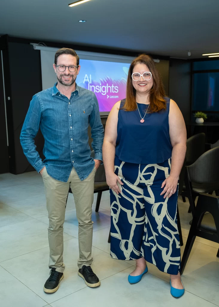 Um homem com camisa jeans e calça bege e uma mulher com óculos de armação branca e calça estampada em pé, lado a lado, sorrindo para a câmera em um auditório. Atrás deles, uma tela de projeção com o texto AI Insights by Lecom.