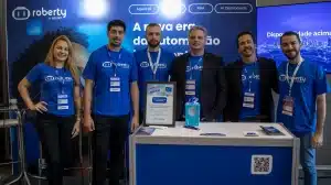 Seis integrantes da equipe Roberty by Lecom, vestindo camisetas azuis da marca, sorriem atrás de um balcão em um estande de evento corporativo de tecnologia.