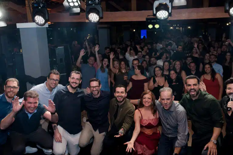 Colaboradores da Lecom Tecnologia reunidos em evento de comemoração, sorrindo, acenando e celebrando juntos sob iluminação de palco.
