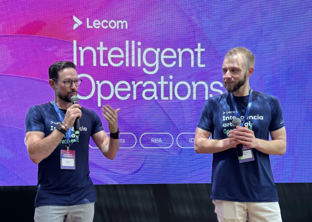 Palestrantes Tiago Amor e Douglas Zilio em apresentação no palco do 1º Lecom Intelligent Operations em São Paulo, diante de uma grande tela de LED com o logotipo da marca e o título do evento.