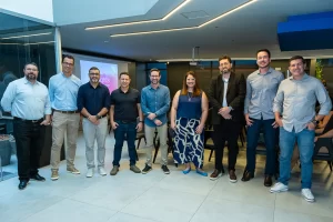 Nove profissionais (oito homens e uma mulher) estão alinhados lado a lado, sorrindo para a câmera em um auditório corporativo moderno e bem iluminado. A mulher (a mesma da imagem anterior, vestindo regata azul-marinho e calça ampla estampada) está posicionada no centro do grupo.
