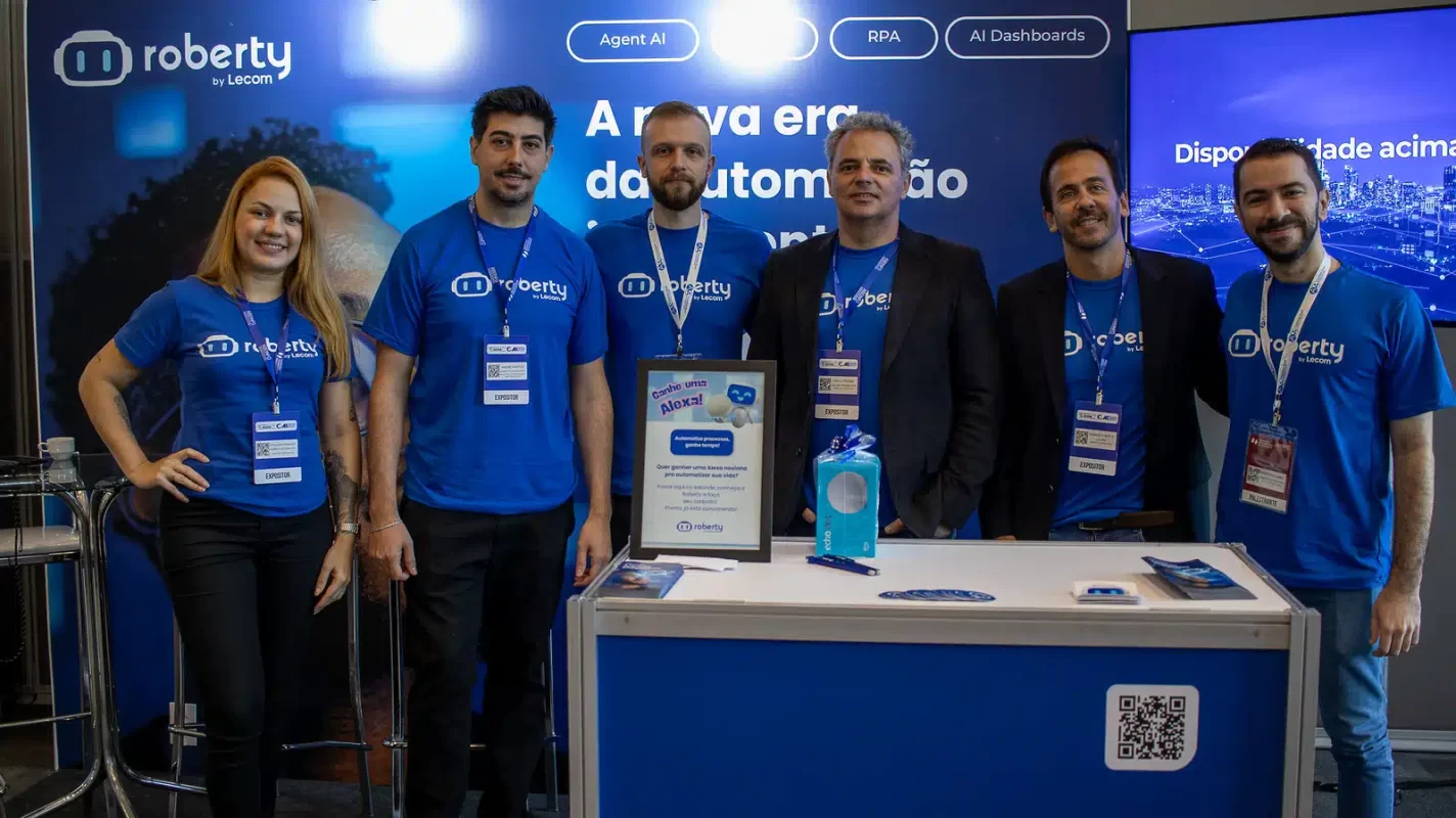 Seis integrantes da equipe Roberty by Lecom, vestindo camisetas azuis da marca, sorriem atrás de um balcão em um estande de evento corporativo de tecnologia.