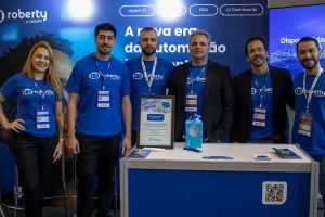 Seis integrantes da equipe Roberty by Lecom, vestindo camisetas azuis da marca, sorriem atrás de um balcão em um estande de evento corporativo de tecnologia.